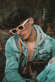 A person wearing white-rimmed sunglasses and a denim jacket layered over a gray hoodie is seated in an outdoor setting. They are carrying a designer bag with a distinct monogram pattern. Their hair is styled voluminously, and there is a relaxed yet introspective demeanor.