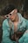 A person wearing white-rimmed sunglasses and a denim jacket layered over a gray hoodie is seated in an outdoor setting. They are carrying a designer bag with a distinct monogram pattern. Their hair is styled voluminously, and there is a relaxed yet introspective demeanor.