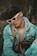 A person wearing white-rimmed sunglasses and a denim jacket layered over a gray hoodie is seated in an outdoor setting. They are carrying a designer bag with a distinct monogram pattern. Their hair is styled voluminously, and there is a relaxed yet introspective demeanor.