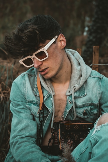 A person wearing white-rimmed sunglasses and a denim jacket layered over a gray hoodie is seated in an outdoor setting. They are carrying a designer bag with a distinct monogram pattern. Their hair is styled voluminously, and there is a relaxed yet introspective demeanor.