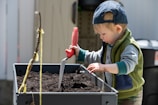 A young child wearing a baseball cap and a green and gray outfit is engaged in gardening. The child is holding a small gardening tool with a red handle and digging into a planter filled with soil. The child appears focused and has dirt on their hands.