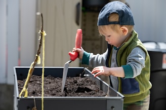 A young child wearing a baseball cap and a green and gray outfit is engaged in gardening. The child is holding a small gardening tool with a red handle and digging into a planter filled with soil. The child appears focused and has dirt on their hands.