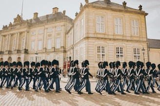 A group of uniformed guards, wearing tall black hats and dark uniforms with blue trousers, march in formation on a cobblestone surface. The background features a historic, elegant building with stone walls and numerous windows, suggesting a ceremonial or official setting.