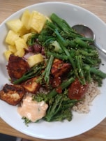 A white bowl contains a colorful and healthy meal featuring bright yellow pineapple chunks, vibrant green broccolini and green beans, grilled pieces of tofu glazed with a red sauce, white rice, and a dollop of creamy pinkish-orange sauce. A metal spoon is resting on the edge of the bowl.