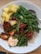 A white bowl contains a colorful and healthy meal featuring bright yellow pineapple chunks, vibrant green broccolini and green beans, grilled pieces of tofu glazed with a red sauce, white rice, and a dollop of creamy pinkish-orange sauce. A metal spoon is resting on the edge of the bowl.