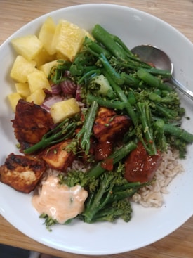 A white bowl contains a colorful and healthy meal featuring bright yellow pineapple chunks, vibrant green broccolini and green beans, grilled pieces of tofu glazed with a red sauce, white rice, and a dollop of creamy pinkish-orange sauce. A metal spoon is resting on the edge of the bowl.