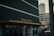 A modern building with a sign reading 'GOOD HOTEL' in large yellow letters on a dark facade. The structure features a minimalist design and is located in an urban setting, surrounded by other tall buildings and a parking area. The overall atmosphere is urban and contemporary.