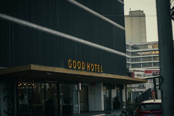 A modern building with a sign reading 'GOOD HOTEL' in large yellow letters on a dark facade. The structure features a minimalist design and is located in an urban setting, surrounded by other tall buildings and a parking area. The overall atmosphere is urban and contemporary.