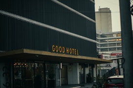A modern building with a sign reading 'GOOD HOTEL' in large yellow letters on a dark facade. The structure features a minimalist design and is located in an urban setting, surrounded by other tall buildings and a parking area. The overall atmosphere is urban and contemporary.