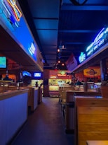 A dimly lit indoor space features wooden booths, with neon signs advertising various brands adorning the walls. The space gives a sense of a lively bar or casual dining restaurant, with visible ceiling lights hanging above.