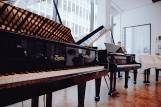 Several grand pianos are elegantly displayed in a well-lit showroom with large windows. The room features wooden floors and feels spacious, with reflections visible on the piano surfaces. A person is seen in the background, possibly interacting with one of the pianos.
