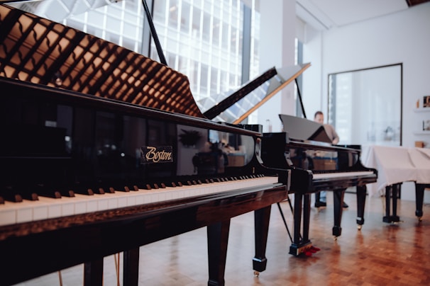 Several grand pianos are elegantly displayed in a well-lit showroom with large windows. The room features wooden floors and feels spacious, with reflections visible on the piano surfaces. A person is seen in the background, possibly interacting with one of the pianos.