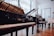 Several grand pianos are elegantly displayed in a well-lit showroom with large windows. The room features wooden floors and feels spacious, with reflections visible on the piano surfaces. A person is seen in the background, possibly interacting with one of the pianos.