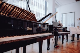 Several grand pianos are elegantly displayed in a well-lit showroom with large windows. The room features wooden floors and feels spacious, with reflections visible on the piano surfaces. A person is seen in the background, possibly interacting with one of the pianos.