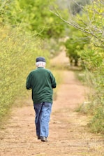 An elderly person is walking away on a dirt path surrounded by lush greenery. The individual is wearing a green jacket, blue jeans, and a dark cap. Branches from trees on either side extend over the path.