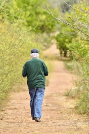 An elderly person is walking away on a dirt path surrounded by lush greenery. The individual is wearing a green jacket, blue jeans, and a dark cap. Branches from trees on either side extend over the path.