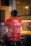 A person wearing a red shirt with the words 'Proudly Independent' is inside a shop, viewed through a window. The interior is softly blurred with warm lighting from the counter display, possibly showing trays of food.