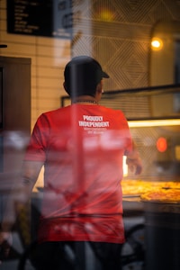 A person wearing a red shirt with the words 'Proudly Independent' is inside a shop, viewed through a window. The interior is softly blurred with warm lighting from the counter display, possibly showing trays of food.
