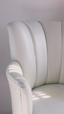 A close-up view of a plush, white upholstered chair with soft, curved armrests. The chair is illuminated by natural light, casting gentle shadows on the surface. The minimalist design and smooth texture are highlighted in the bright setting.