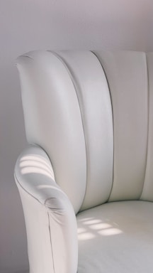 A close-up view of a plush, white upholstered chair with soft, curved armrests. The chair is illuminated by natural light, casting gentle shadows on the surface. The minimalist design and smooth texture are highlighted in the bright setting.