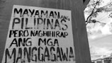 A wall with a poster in black and white text written in the Filipino language. In the background, there is an outdoor scene with utility lines, a tree, and a partly cloudy sky.
