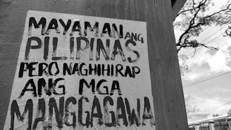 A wall with a poster in black and white text written in the Filipino language. In the background, there is an outdoor scene with utility lines, a tree, and a partly cloudy sky.