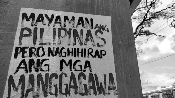 A wall with a poster in black and white text written in the Filipino language. In the background, there is an outdoor scene with utility lines, a tree, and a partly cloudy sky.