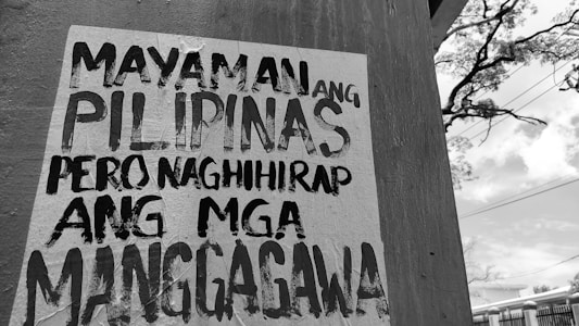 A wall with a poster in black and white text written in the Filipino language. In the background, there is an outdoor scene with utility lines, a tree, and a partly cloudy sky.