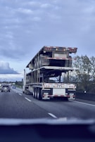 A flatbed truck is transporting another flatbed trailer on a highway. The sky is overcast, and other vehicles are present on the road. The surrounding landscape includes trees and signs. The perspective is from another vehicle.