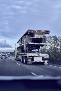 A flatbed truck is transporting another flatbed trailer on a highway. The sky is overcast, and other vehicles are present on the road. The surrounding landscape includes trees and signs. The perspective is from another vehicle.