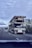 A flatbed truck is transporting another flatbed trailer on a highway. The sky is overcast, and other vehicles are present on the road. The surrounding landscape includes trees and signs. The perspective is from another vehicle.