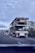 A flatbed truck is transporting another flatbed trailer on a highway. The sky is overcast, and other vehicles are present on the road. The surrounding landscape includes trees and signs. The perspective is from another vehicle.