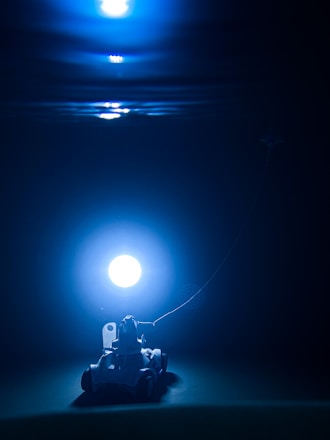 A small vehicle with wheels is positioned underwater, tethered to a glowing light source. The scene is bathed in a deep blue hue, creating an ethereal and mysterious ambiance.