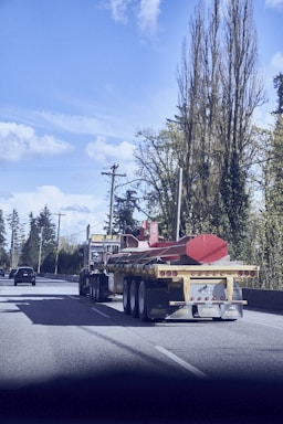 A large flatbed truck is driving down a highway, carrying a long, red industrial component. The sky is bright with scattered clouds, and tall trees line the side of the road. Several cars are also visible on the highway.