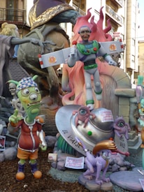 A whimsical scene featuring colorful sculptures with a playful and cartoonish style. A central figure resembles a parody of a superhero with comical facial features, holding a microphone. Surrounding the main character are other fantastical creatures, including an alien with one eye and a humorous expression, and a purple animal with exaggerated features. An object resembling a flying saucer is also present, supporting two small one-eyed creatures. The background shows more of the sculptural display with additional characters and decorative elements.