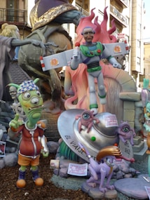 A whimsical scene featuring colorful sculptures with a playful and cartoonish style. A central figure resembles a parody of a superhero with comical facial features, holding a microphone. Surrounding the main character are other fantastical creatures, including an alien with one eye and a humorous expression, and a purple animal with exaggerated features. An object resembling a flying saucer is also present, supporting two small one-eyed creatures. The background shows more of the sculptural display with additional characters and decorative elements.