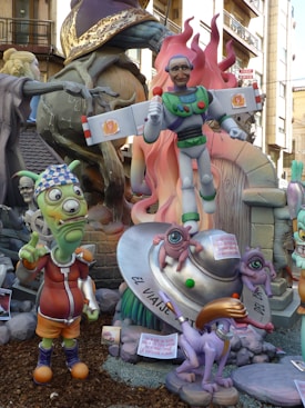 A whimsical scene featuring colorful sculptures with a playful and cartoonish style. A central figure resembles a parody of a superhero with comical facial features, holding a microphone. Surrounding the main character are other fantastical creatures, including an alien with one eye and a humorous expression, and a purple animal with exaggerated features. An object resembling a flying saucer is also present, supporting two small one-eyed creatures. The background shows more of the sculptural display with additional characters and decorative elements.