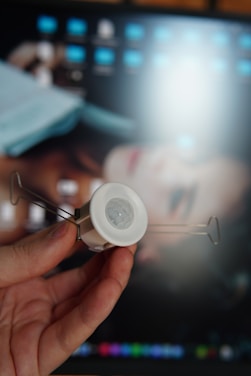 A hand is holding a small, white electronic device with a lens in the center, likely a sensor, which is flanked by metal arms. In the background, there is a blurry image of a computer screen displaying various icons and a digital image of a face.