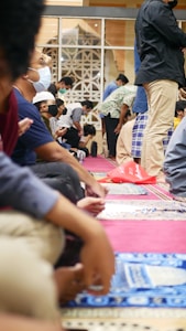 Individuals are seated on the floor in a setting that suggests a communal or religious gathering. They appear to be focused and engaged, with most of them wearing face masks. Some are wearing head coverings, and the atmosphere suggests quiet attentiveness. The decor includes patterned carpets and architectural elements like arches in the background.