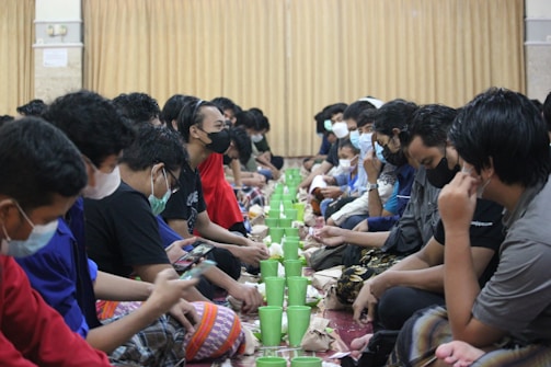 A group of people sitting on the floor in two lines facing each other, many of whom are wearing face masks. In front of them, there are green plastic cups and bundled packages, suggesting a communal meal setting. The background consists of beige curtains, adding a warm tone to the environment.