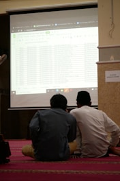 Two individuals are seated on a carpeted floor facing a projected screen displaying a spreadsheet. The projection is detailed with various data columns, and the environment suggests a formal or educational setting. A sign on the wall reads 'DILARANG MEMBUKA MASKER'.