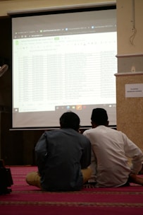 Two individuals are seated on a carpeted floor facing a projected screen displaying a spreadsheet. The projection is detailed with various data columns, and the environment suggests a formal or educational setting. A sign on the wall reads 'DILARANG MEMBUKA MASKER'.