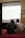 Two individuals are seated on a carpeted floor facing a projected screen displaying a spreadsheet. The projection is detailed with various data columns, and the environment suggests a formal or educational setting. A sign on the wall reads 'DILARANG MEMBUKA MASKER'.