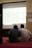 Two individuals are seated on a carpeted floor facing a projected screen displaying a spreadsheet. The projection is detailed with various data columns, and the environment suggests a formal or educational setting. A sign on the wall reads 'DILARANG MEMBUKA MASKER'.