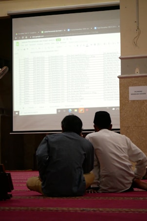 Two individuals are seated on a carpeted floor facing a projected screen displaying a spreadsheet. The projection is detailed with various data columns, and the environment suggests a formal or educational setting. A sign on the wall reads 'DILARANG MEMBUKA MASKER'.