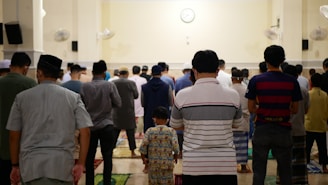 A group of people standing in rows, appearing to be engaged in a communal activity in an indoor setting. The individuals are facing away from the camera, with fans mounted on the walls and a clock visible at the front of the room.