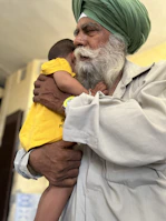 An elderly man with a white beard and a green turban gently holds a young child who is wearing a bright yellow shirt. The man’s expression is calm and tender as he embraces the child, providing a sense of warmth and protection. The background is softly blurred, emphasizing the connection between the two.
