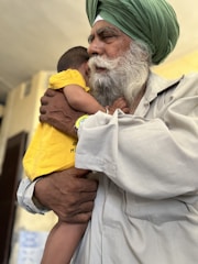 An elderly man with a white beard and a green turban gently holds a young child who is wearing a bright yellow shirt. The man’s expression is calm and tender as he embraces the child, providing a sense of warmth and protection. The background is softly blurred, emphasizing the connection between the two.
