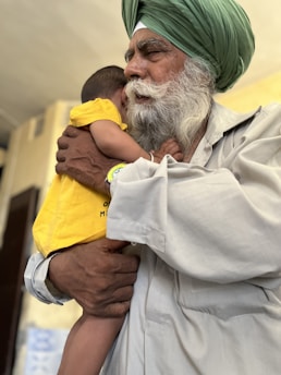 An elderly man with a white beard and a green turban gently holds a young child who is wearing a bright yellow shirt. The man’s expression is calm and tender as he embraces the child, providing a sense of warmth and protection. The background is softly blurred, emphasizing the connection between the two.