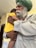 An elderly man with a white beard and a green turban gently holds a young child who is wearing a bright yellow shirt. The man’s expression is calm and tender as he embraces the child, providing a sense of warmth and protection. The background is softly blurred, emphasizing the connection between the two.